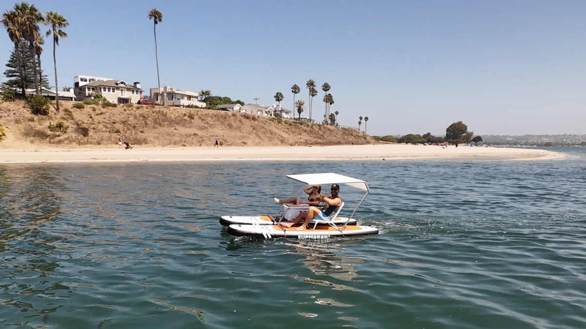 A couple relaxing under the canopy of a Supmaran inflatable catamaran, cruising on calm water near a sandy beach and coastal homes.